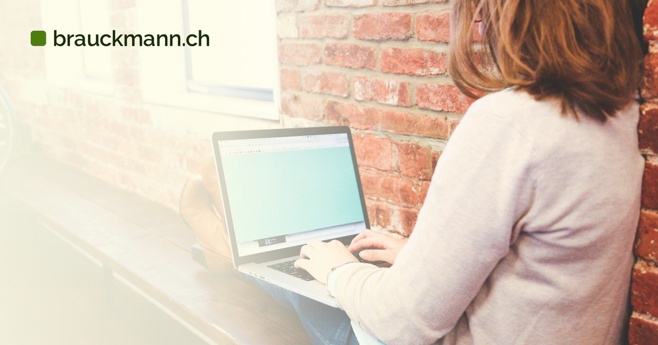 Frau sitzt vor einer Backsteinwand und tippt auf einem Laptop mit hellem Bildschirm