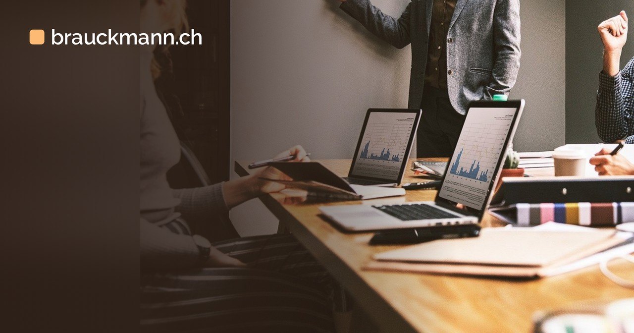 Team am Holztisch im Besprechungsraum, auf den Laptops sind Balkendiagramme zu sehen, im Hintergrund präsentiert jemand